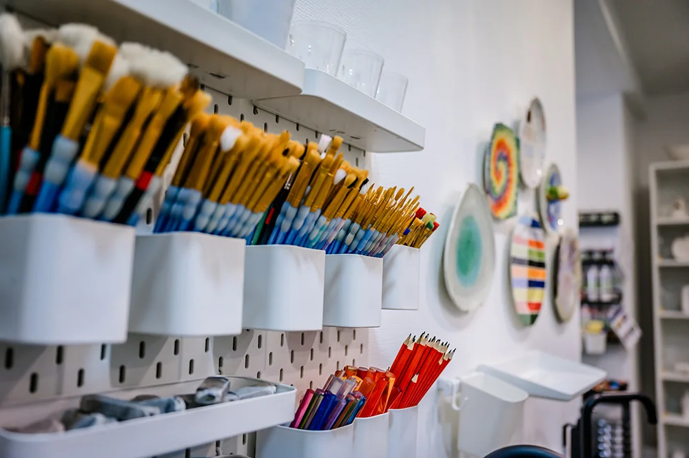 An einer weißen Wand bei Farbenfleck in Aachen befinden sich Regale mit Gläsern, die mit Pinseln und Buntstiften gefüllt sind. Im Hintergrund an der angrenzenden Wand sind dekorative bemalte Teller ausgestellt.